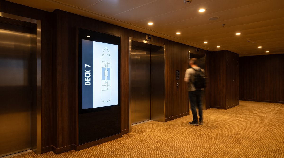 A lit portrait screen beside cruise ship lift doors showing a deck plan