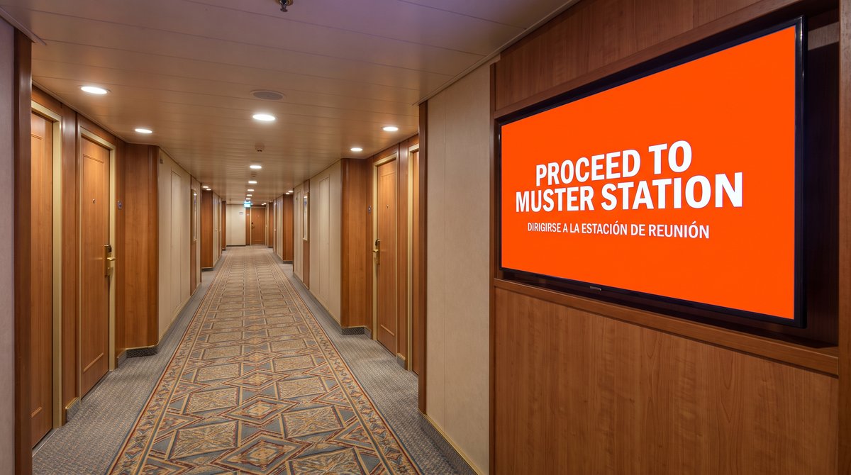 A screen in a cruise ship corridor showing a muster instruction in two languages